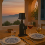 Akku-Tischleuchte in Form einer Flasche auf einem gedeckten Tisch bei Sonnenuntergang, seitliche Perspektive