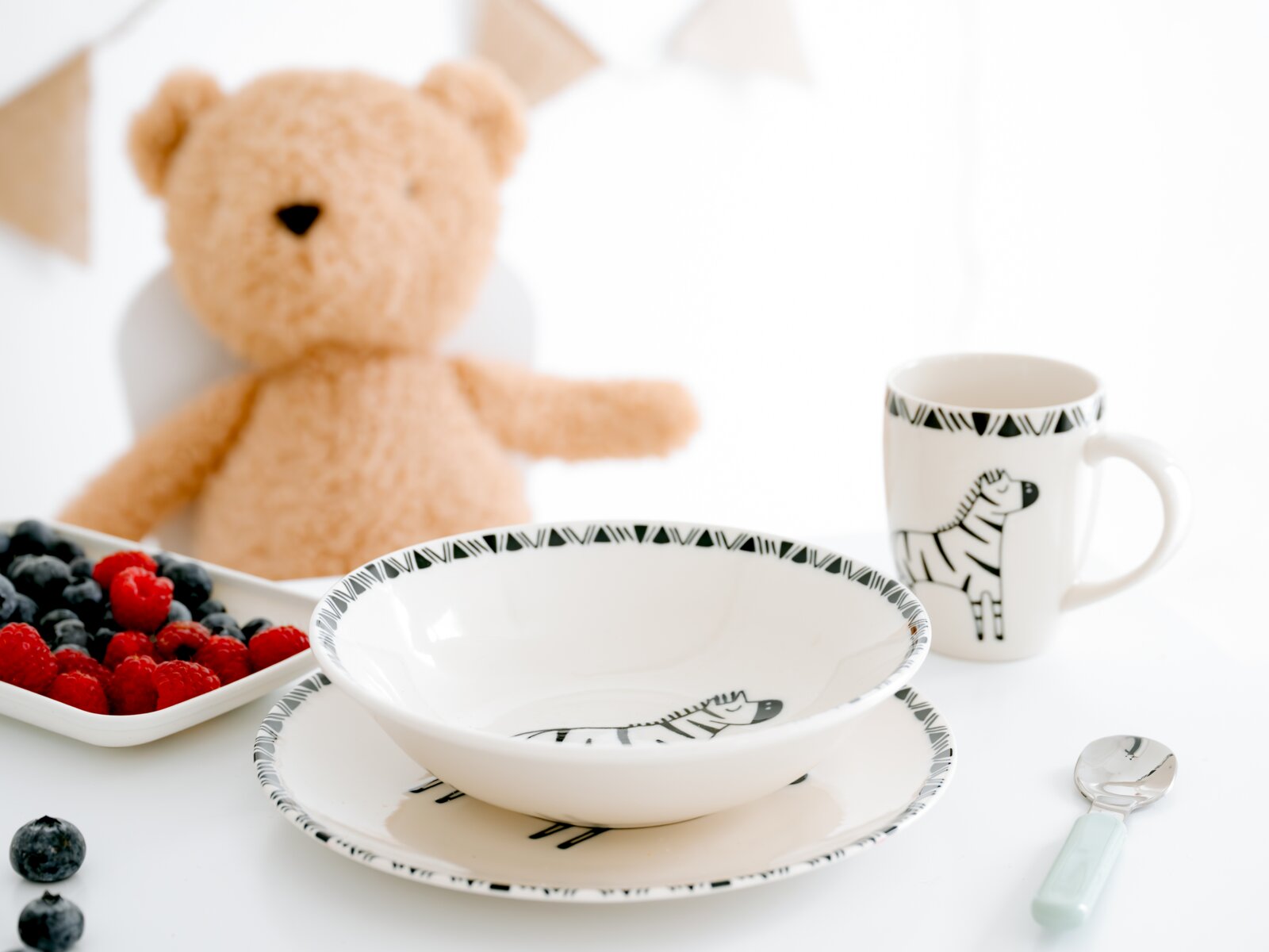 Weißes Kinder-Geschirrset mit schwarzen Zebramotiven, bestehend aus Schale, Teller und Henkelbecher, aus leicht erhöhter Frontalperspektive arrangiert mit Beeren, Löffel und Teddybär.