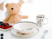 Weißes Kinder-Geschirrset mit schwarzen Zebramotiven, bestehend aus Schale, Teller und Henkelbecher, aus leicht erhöhter Frontalperspektive arrangiert mit Beeren, Löffel und Teddybär.