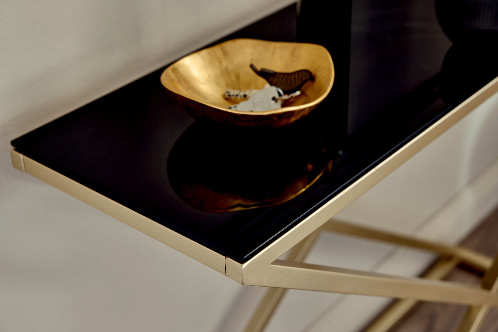 Nahaufnahme einer eleganten Konsole mit schwarzer Glasoberfläche und goldfarbenem Gestell, darauf eine goldene Schale mit Schlüsseln.
