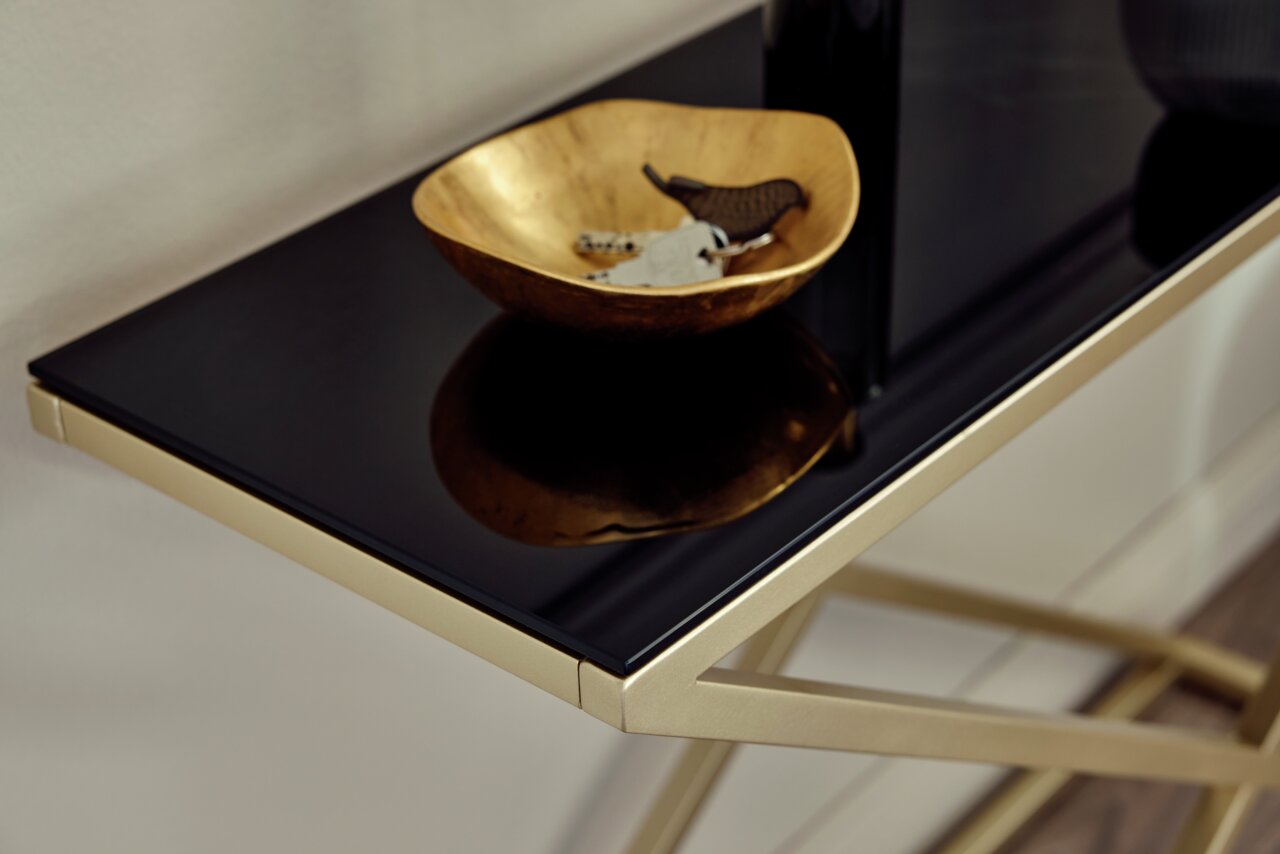 Close-up van een elegante console met een zwart glazen blad en een goudkleurig onderstel, met daarop een gouden schaal met sleutels.