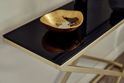 Nahaufnahme einer eleganten Konsole mit schwarzer Glasoberfläche und goldfarbenem Gestell, darauf eine goldene Schale mit Schlüsseln.
