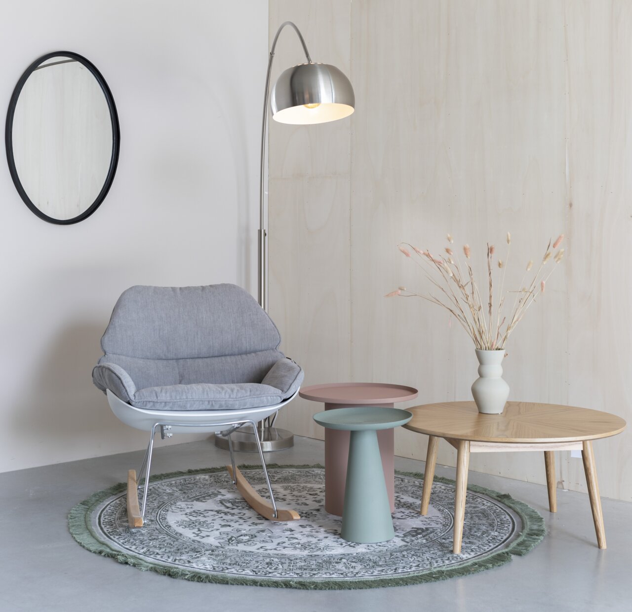 Moderne schommelstoel in grijs, geplaatst in een elegante woonkamer met bijzettafels en een staande lamp, gezien vanuit de zijkant.