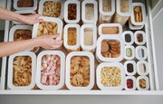 Vogelperspectief op een lade met verschillende witte voorraadbakjes, gevuld met etenswaren zoals pasta, koekjes en popcorn.