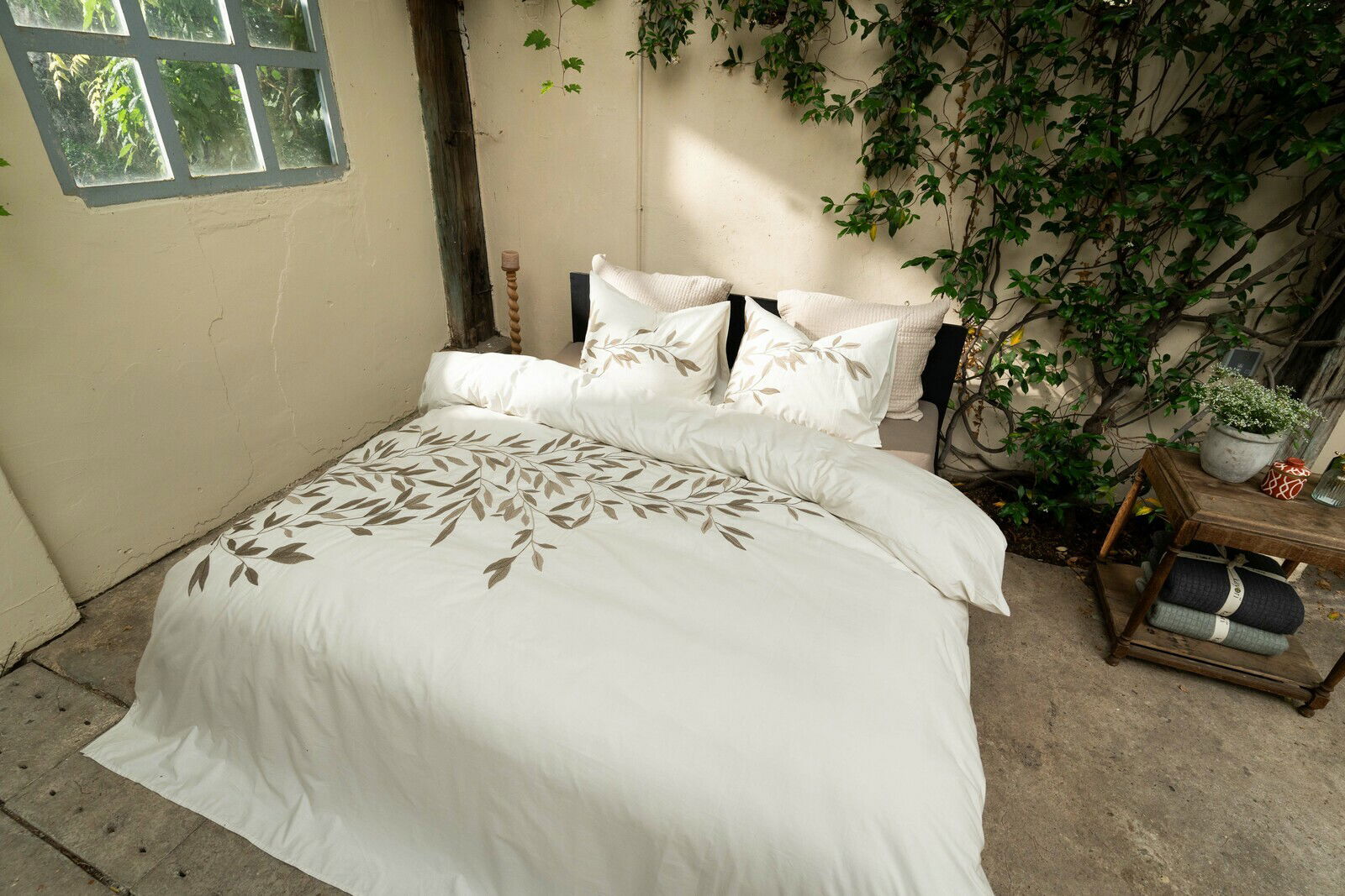 Bettwäsche-Set in einem gemütlichen Raum mit Pflanzen, bestehend aus einem Bettbezug und Kissenbezügen mit einem eleganten Blattmuster, fotografiert aus einer leicht erhöhten Perspektive.