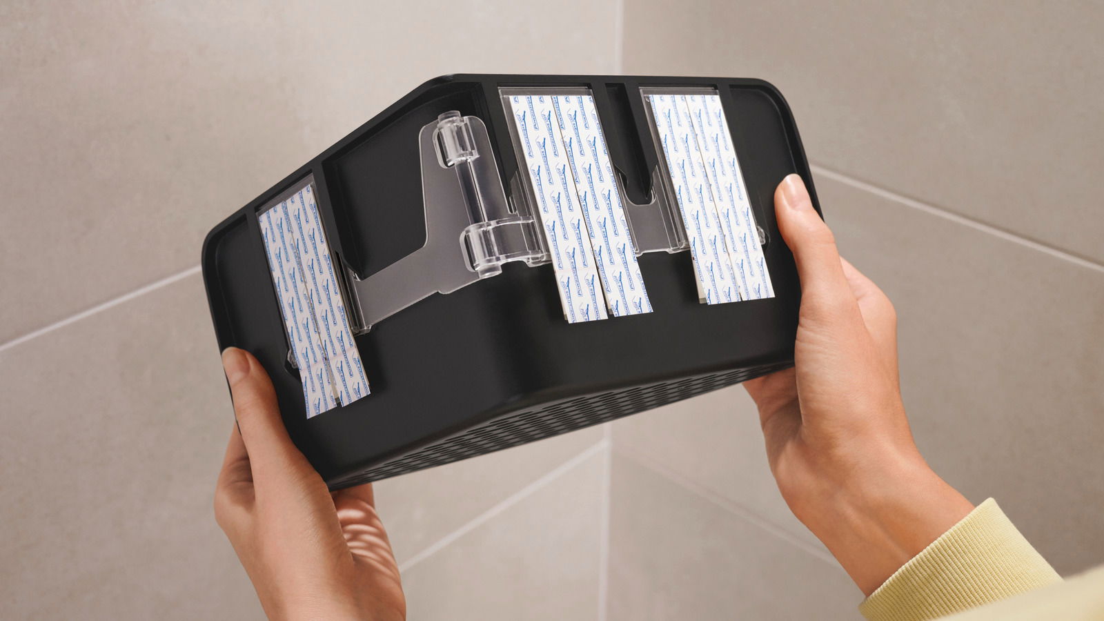 Nahaufnahme eines schwarzen Eckduschkorbs von unten, gehalten von zwei Händen, mit sichtbaren Klebestreifen zur Befestigung.