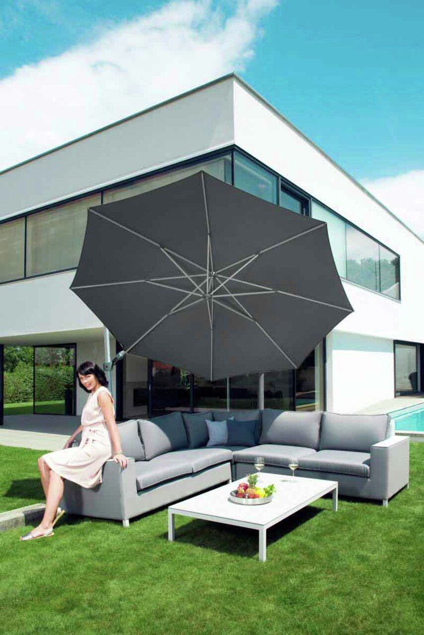 Moderner Pendelschirm über einer eleganten Outdoor-Sitzgruppe mit einer Frau, die auf dem Sofa sitzt, vor einem modernen Haus. Perspektive von schräg vorne.