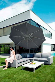 Moderner Pendelschirm über einer eleganten Outdoor-Sitzgruppe mit einer Frau, die auf dem Sofa sitzt, vor einem modernen Haus. Perspektive von schräg vorne.