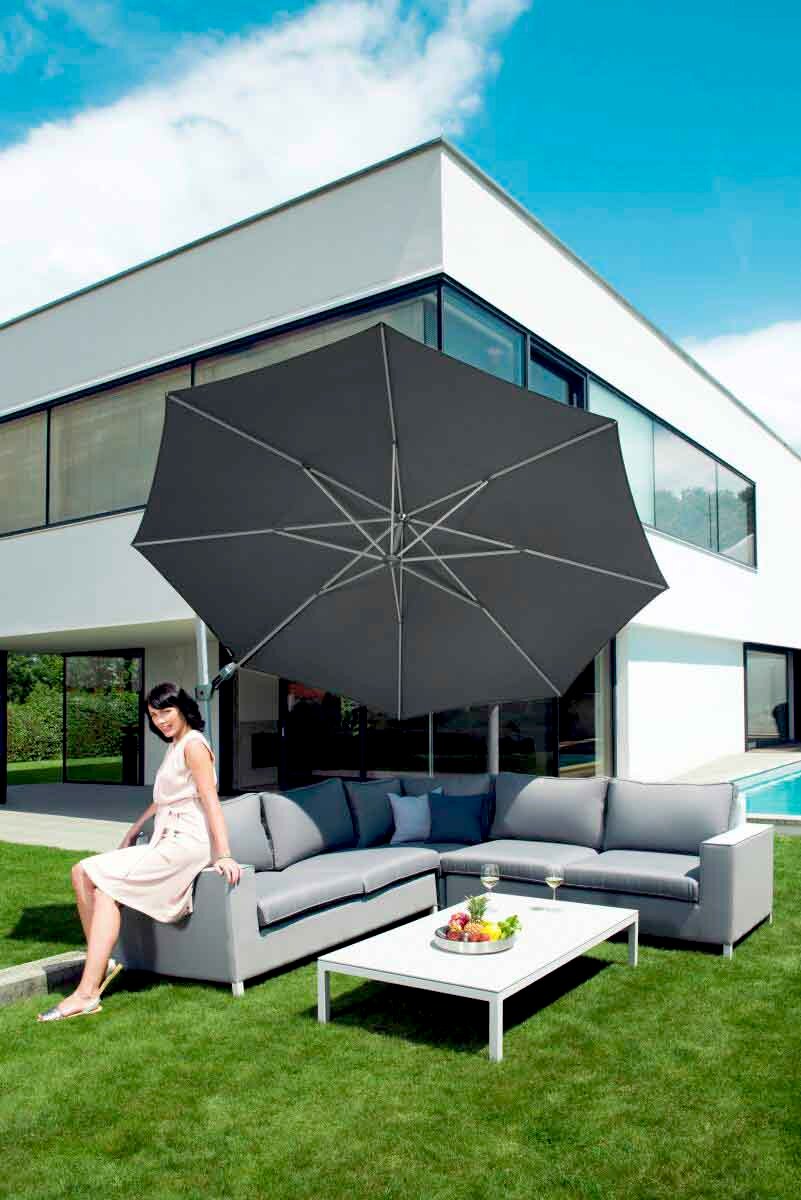 Moderner Pendelschirm über einer eleganten Outdoor-Sitzgruppe mit einer Frau, die auf dem Sofa sitzt, vor einem modernen Haus. Perspektive von schräg vorne.