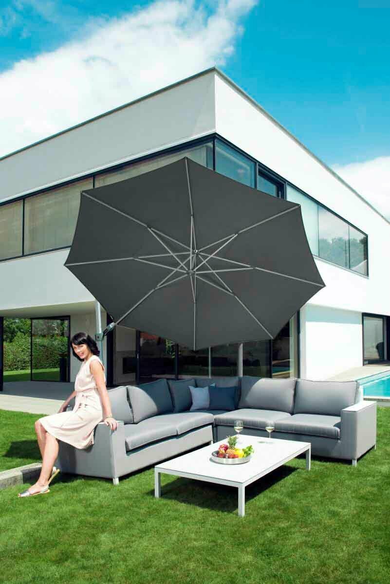 Moderner Pendelschirm über einer eleganten Outdoor-Sitzgruppe mit einer Frau, die auf dem Sofa sitzt, vor einem modernen Haus. Perspektive von schräg vorne.