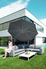Moderner Pendelschirm über einer eleganten Outdoor-Sitzgruppe mit einer Frau, die auf dem Sofa sitzt, vor einem modernen Haus. Perspektive von schräg vorne.