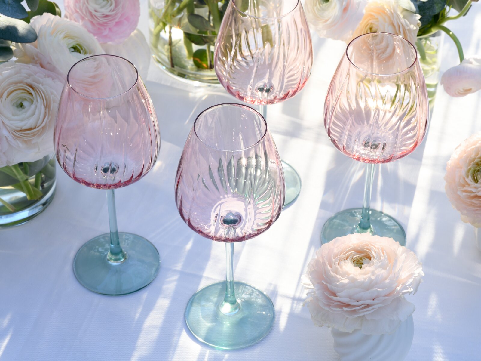 Vier strukturierte Weingläser aus roséfarbenem Glas mit grünlichem Stiel und Fuß auf weißer Tischdecke, dekoriert mit hellen Blumen; Perspektive: leicht von oben in schräger Ansicht.