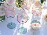 Vier strukturierte Weingläser aus roséfarbenem Glas mit grünlichem Stiel und Fuß auf weißer Tischdecke, dekoriert mit hellen Blumen; Perspektive: leicht von oben in schräger Ansicht.