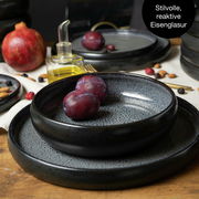 Nahaufnahme eines stilvollen, reaktiven Eisenglasur-Tafelservices in Grau mit Pflaumen und Granatapfel im Hintergrund.