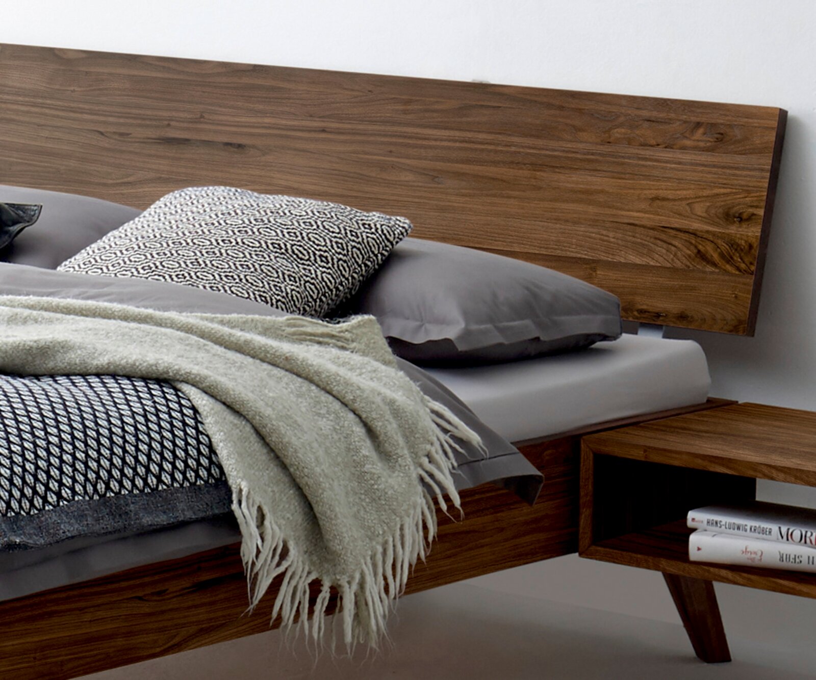 Modernes Bettgestell aus Holz mit Kopfteil, seitliche Perspektive, dekoriert mit grauen Kissen und Decke.
