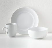 Weißes, minimalistisches Geschirr-Set mit großem Teller, Schale und Henkelbecher auf hellem Hintergrund, frontal in leichter Seitenansicht fotografiert