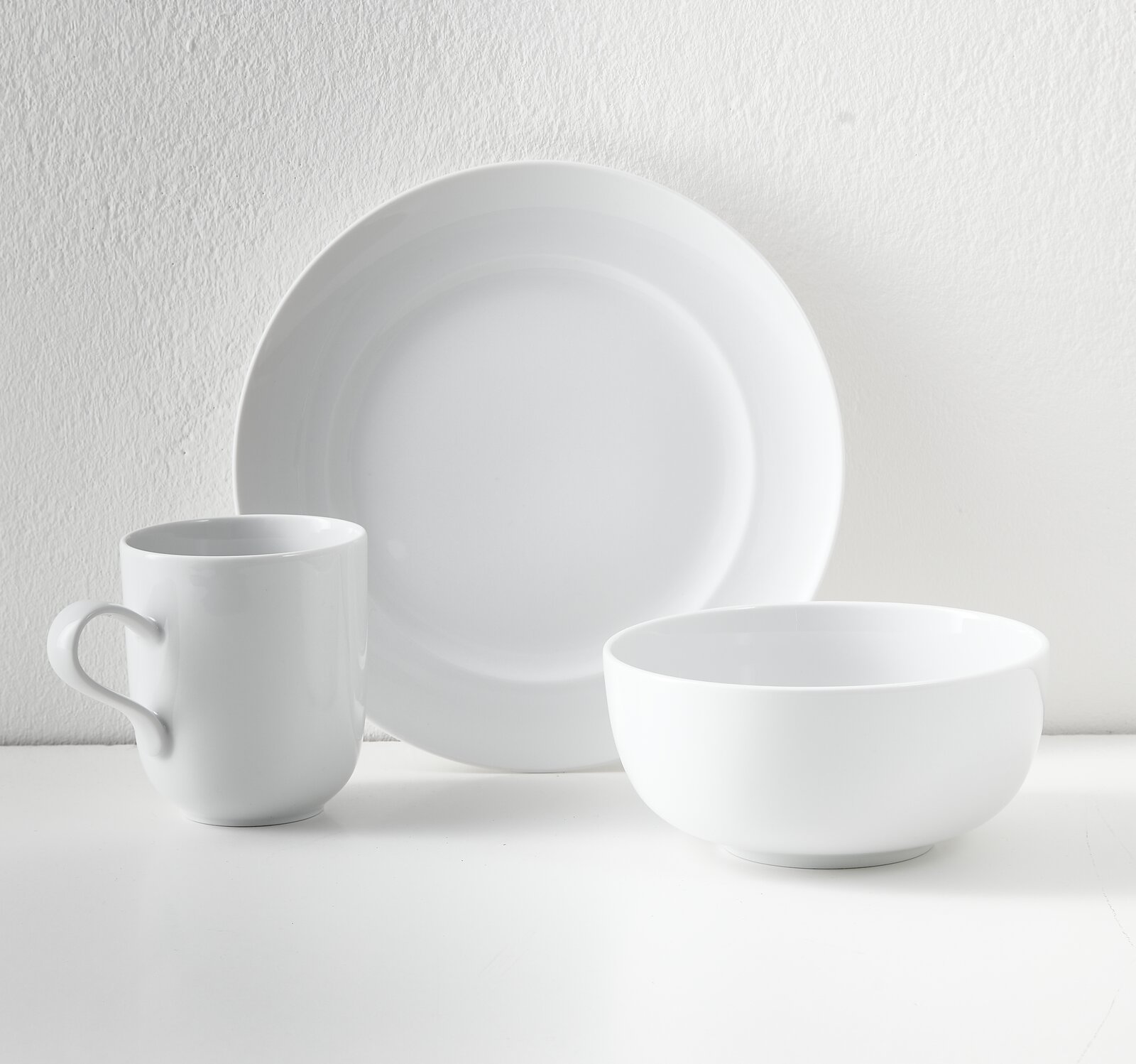 Weißes, minimalistisches Geschirr-Set mit großem Teller, Schale und Henkelbecher auf hellem Hintergrund, frontal in leichter Seitenansicht fotografiert