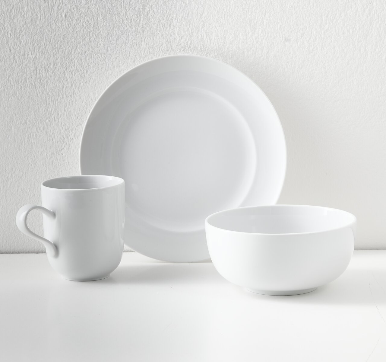 Weißes, minimalistisches Geschirr-Set mit großem Teller, Schale und Henkelbecher auf hellem Hintergrund, frontal in leichter Seitenansicht fotografiert
