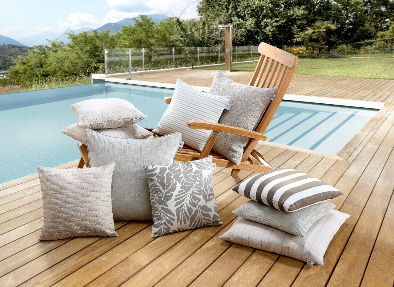 Linneweber Outdoor Kissen COMFORT Outdoor-Kissen in verschiedenen Designs und Größen auf einer Holzterrasse neben einem Pool, mit einem Liegestuhl im Hintergrund.