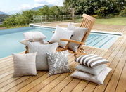 Outdoor-Kissen in verschiedenen Designs und Größen auf einer Holzterrasse neben einem Pool, mit einem Liegestuhl im Hintergrund.