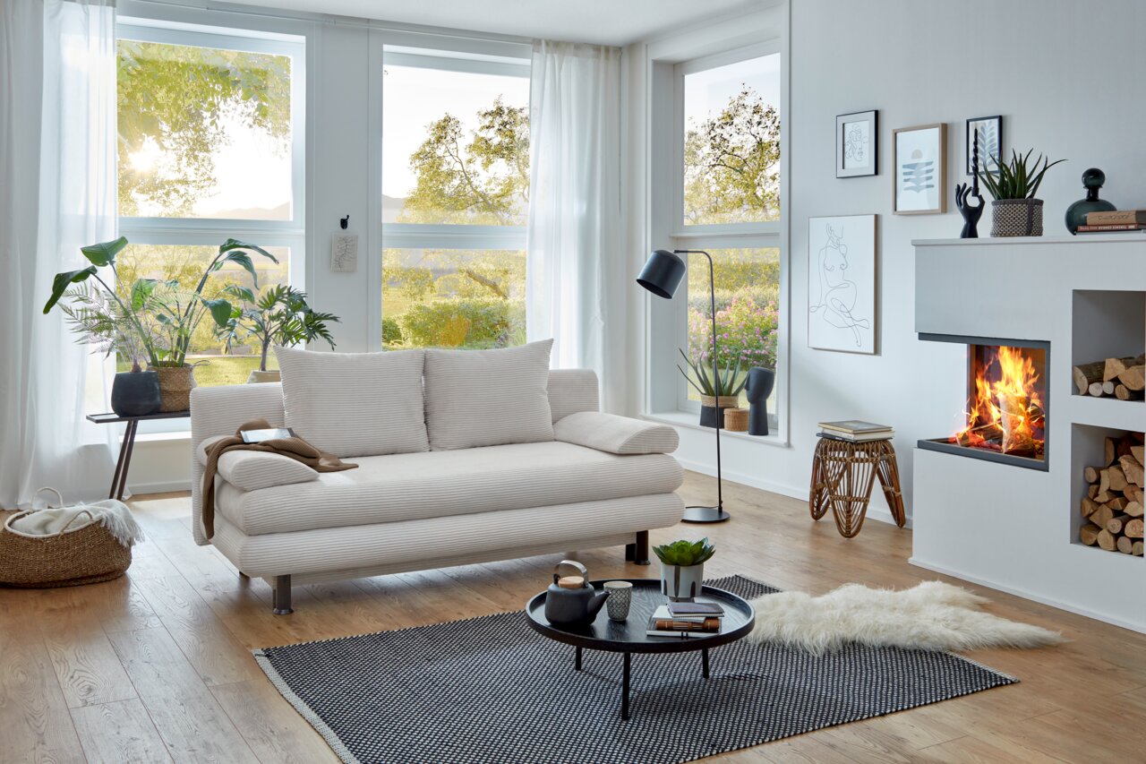 Modernes Funktionssofa in einem hellen Wohnzimmer mit großen Fenstern, Pflanzen und einem Kamin, seitliche Perspektive.