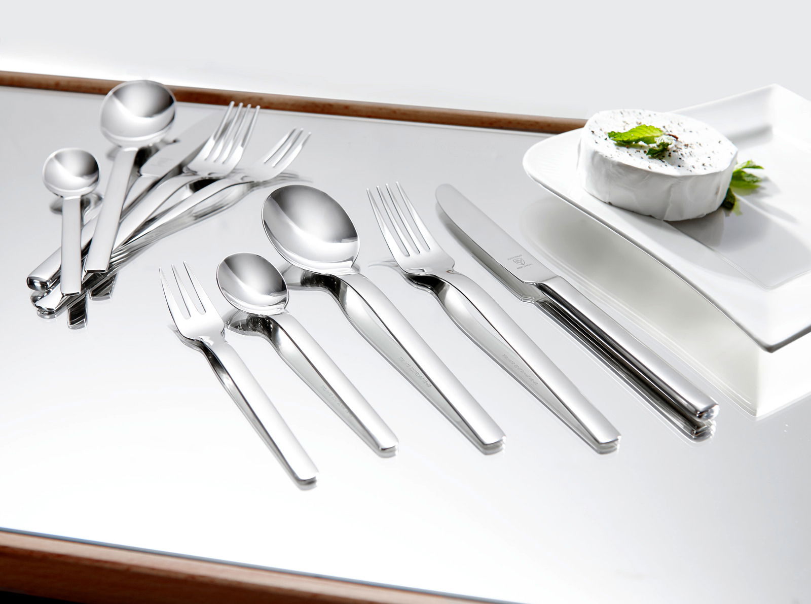 Elegantes 60-teiliges Besteckset aus Edelstahl, von oben betrachtet, auf einem glänzenden Tisch neben einem Teller mit Käse.