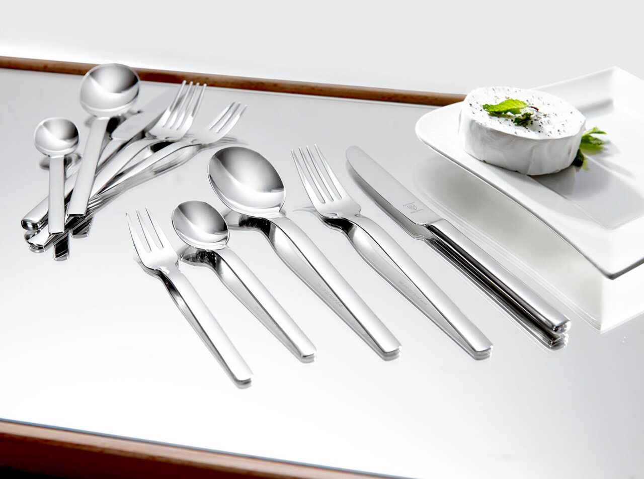 Elegantes 60-teiliges Besteckset aus Edelstahl, von oben betrachtet, auf einem glänzenden Tisch neben einem Teller mit Käse.