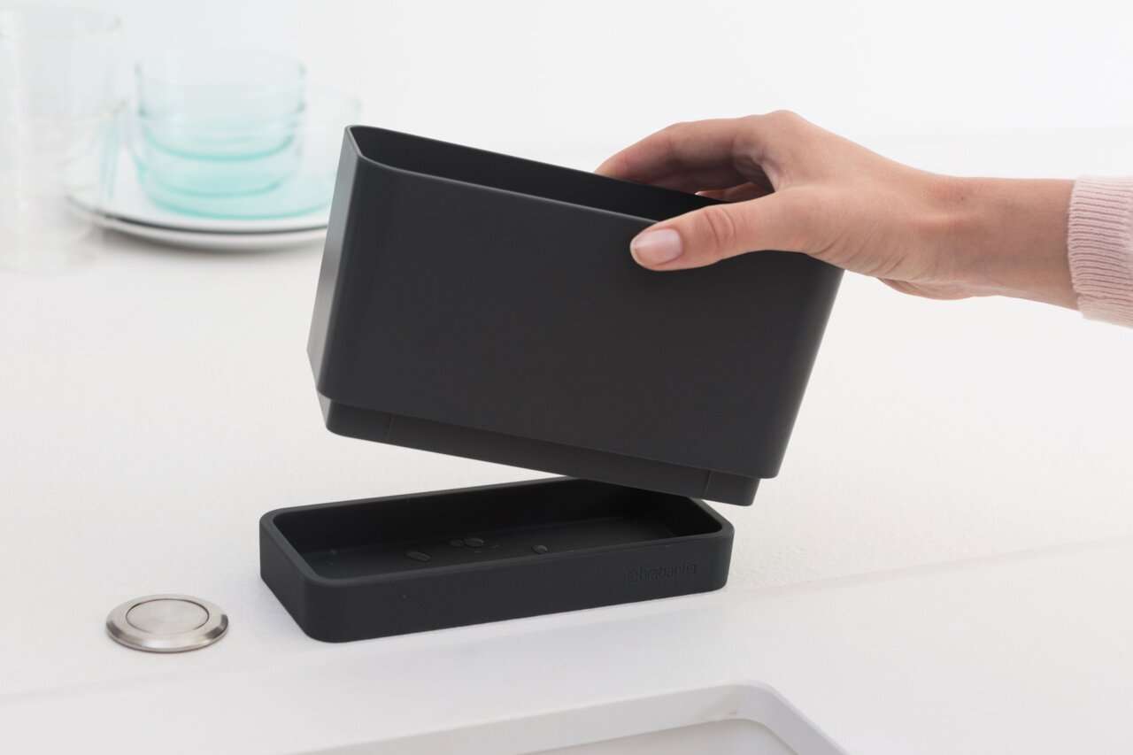 Hand hält einen schwarzen Brabantia Spül-Organizer über einer Abtropfschale in einer Küchenumgebung, seitliche Perspektive.