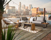 Moderne Gartenlounge auf einer Dachterrasse mit Blick auf die Stadt, bestehend aus einem grauen Sofa mit Kissen, einem Beistelltisch aus Holz und Metall, dekoriert mit Pflanzen und Kerzen.