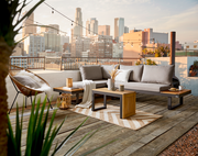 Moderne Gartenlounge auf einer Dachterrasse mit Blick auf die Stadt, bestehend aus einem grauen Sofa mit Kissen, einem Beistelltisch aus Holz und Metall, dekoriert mit Pflanzen und Kerzen.