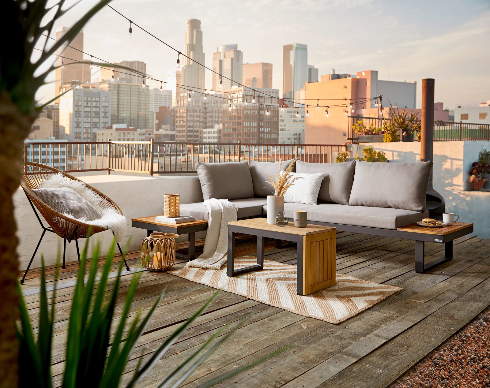 Amatio Tuinlounge GALICIA Moderne Gartenlounge auf einer Dachterrasse mit Blick auf die Stadt, bestehend aus einem grauen Sofa mit Kissen, einem Beistelltisch aus Holz und Metall, dekoriert mit Pflanzen und Kerzen.