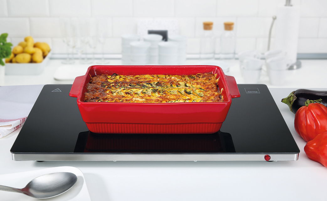 Rote Auflaufform mit gebackenem Gericht auf einer schwarzen Warmhalteplatte in einer modernen Küche, Perspektive von vorne.