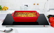 Rote Auflaufform mit gebackenem Gericht auf einer schwarzen Warmhalteplatte in einer modernen Küche, Perspektive von vorne.