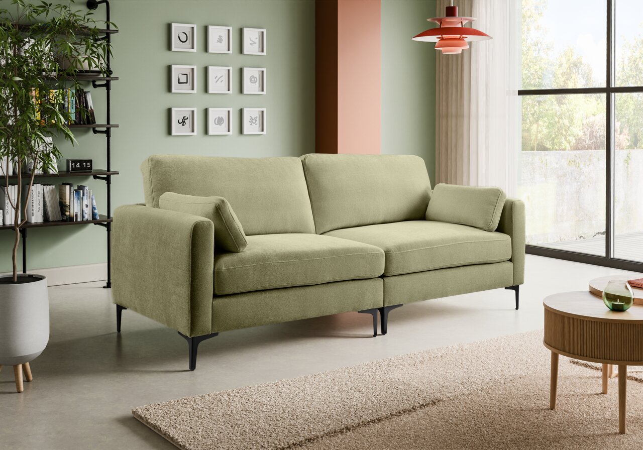 Modernes 2-Sitzer-Sofa in Salbeigrün mit Stoffbezug, breiten Armlehnen, zwei Rückenkissen und schwarzen Metallfüßen im hellen Wohnzimmer – Frontansicht in leichter Schrägperspektive von links.