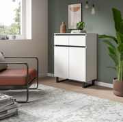 Moderne weiße Kommode mit zwei Schubladen und zwei Türen, seitliche Perspektive, in einem stilvoll eingerichteten Raum mit grüner Wand und Pflanzen.