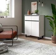 Moderne weiße Kommode mit zwei Schubladen und zwei Türen, seitliche Perspektive, in einem stilvoll eingerichteten Raum mit grüner Wand und Pflanzen.