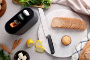 Bovenaanzicht van een wit broodmes met zwarte handgreep op een ronde marmeren plank naast gesneden brood, met op de achtergrond groenten en een zwarte opbergtray met gekleurde dopjes.