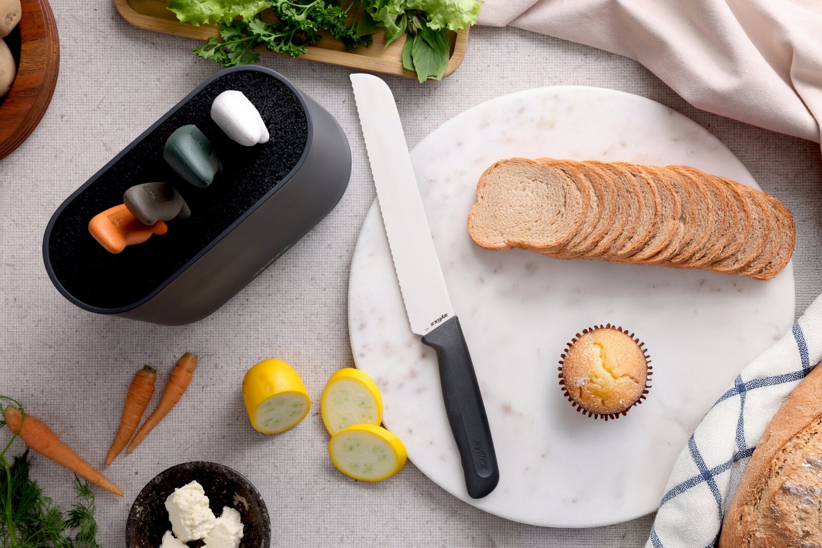 Bovenaanzicht van een wit broodmes met zwarte handgreep op een ronde marmeren plank naast gesneden brood, met op de achtergrond groenten en een zwarte opbergtray met gekleurde dopjes.