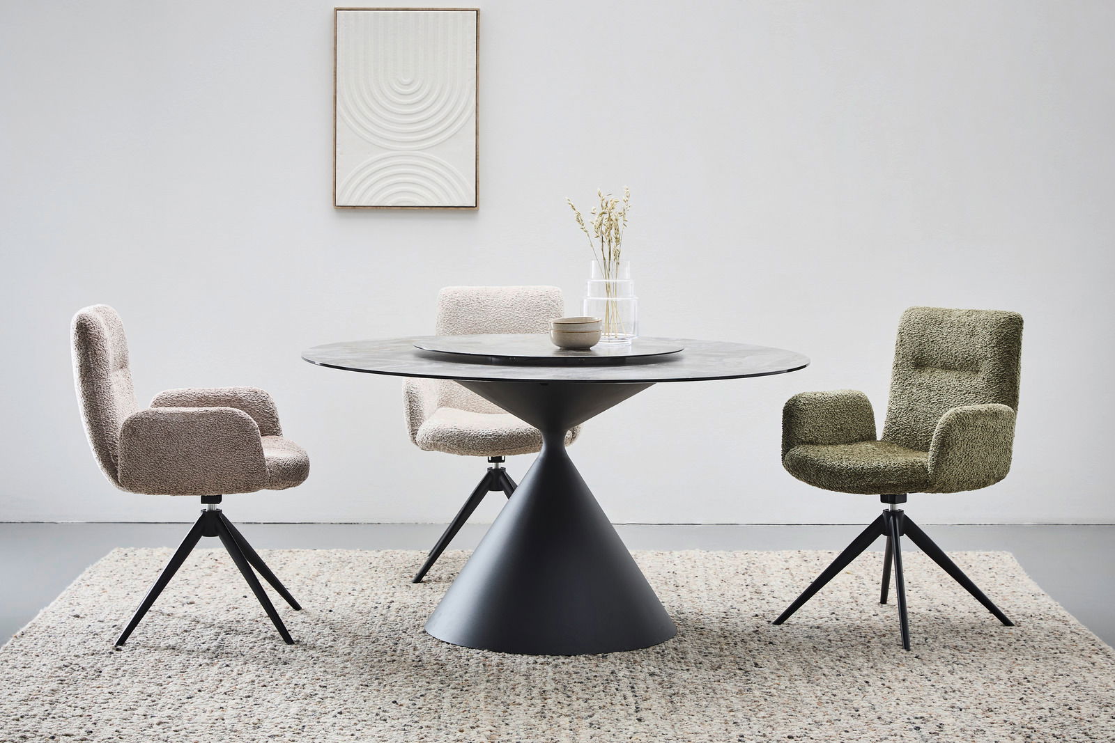 Moderner Esstisch mit rundem, schwarzem Sockel und grauer Tischplatte, umgeben von drei gepolsterten Stühlen in verschiedenen Farben, aus der Frontalperspektive.