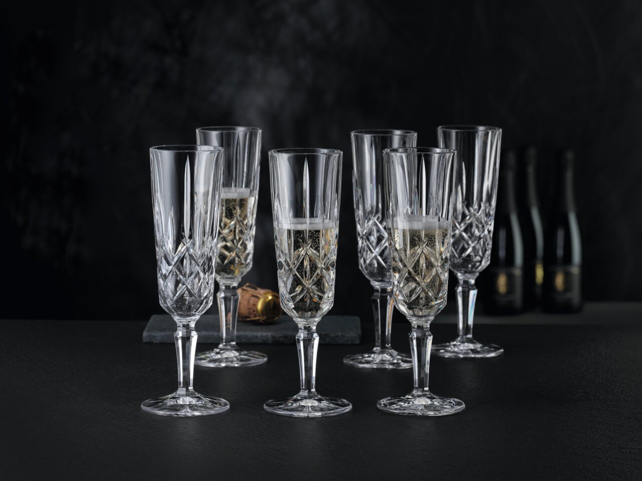 Sechs elegante Champagnergläser aus Kristallglas mit feinem Schliff, gefüllt mit Champagner, auf schwarzem Hintergrund. Perspektive von vorne.