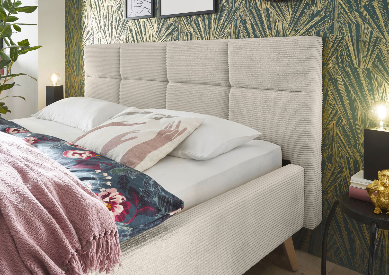 Seitliche Perspektive auf ein modernes Polsterbett mit beigem Kopfteil und dekorativen Kissen, umgeben von stilvoller Tapete und Pflanzen.