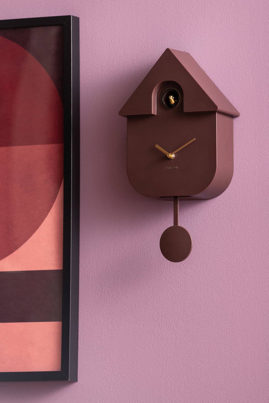 Moderne Wanduhr in Hausform mit Pendel, braun, an einer lila Wand, seitliche Perspektive.