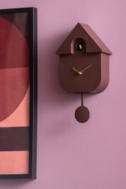 Moderne Wanduhr in Hausform mit Pendel, braun, an einer lila Wand, seitliche Perspektive.