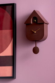 Moderne Wanduhr in Hausform mit Pendel, braun, an einer lila Wand, seitliche Perspektive.