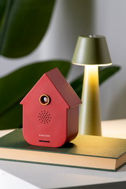 Rote, moderne Soundbox in Hausform, frontal fotografiert, auf einem Buch stehend, mit einer Lampe im Hintergrund.