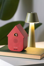 Rote, moderne Soundbox in Hausform, frontal fotografiert, auf einem Buch stehend, mit einer Lampe im Hintergrund.