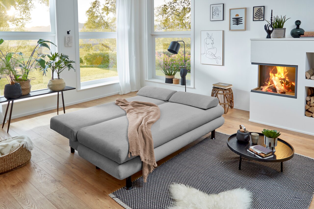 Graues Funktionssofa in einem modernen Wohnzimmer mit großen Fenstern, Pflanzen und Kamin, aus seitlicher Perspektive fotografiert.