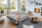 Graues Funktionssofa in einem modernen Wohnzimmer mit großen Fenstern, Pflanzen und Kamin, aus seitlicher Perspektive fotografiert.