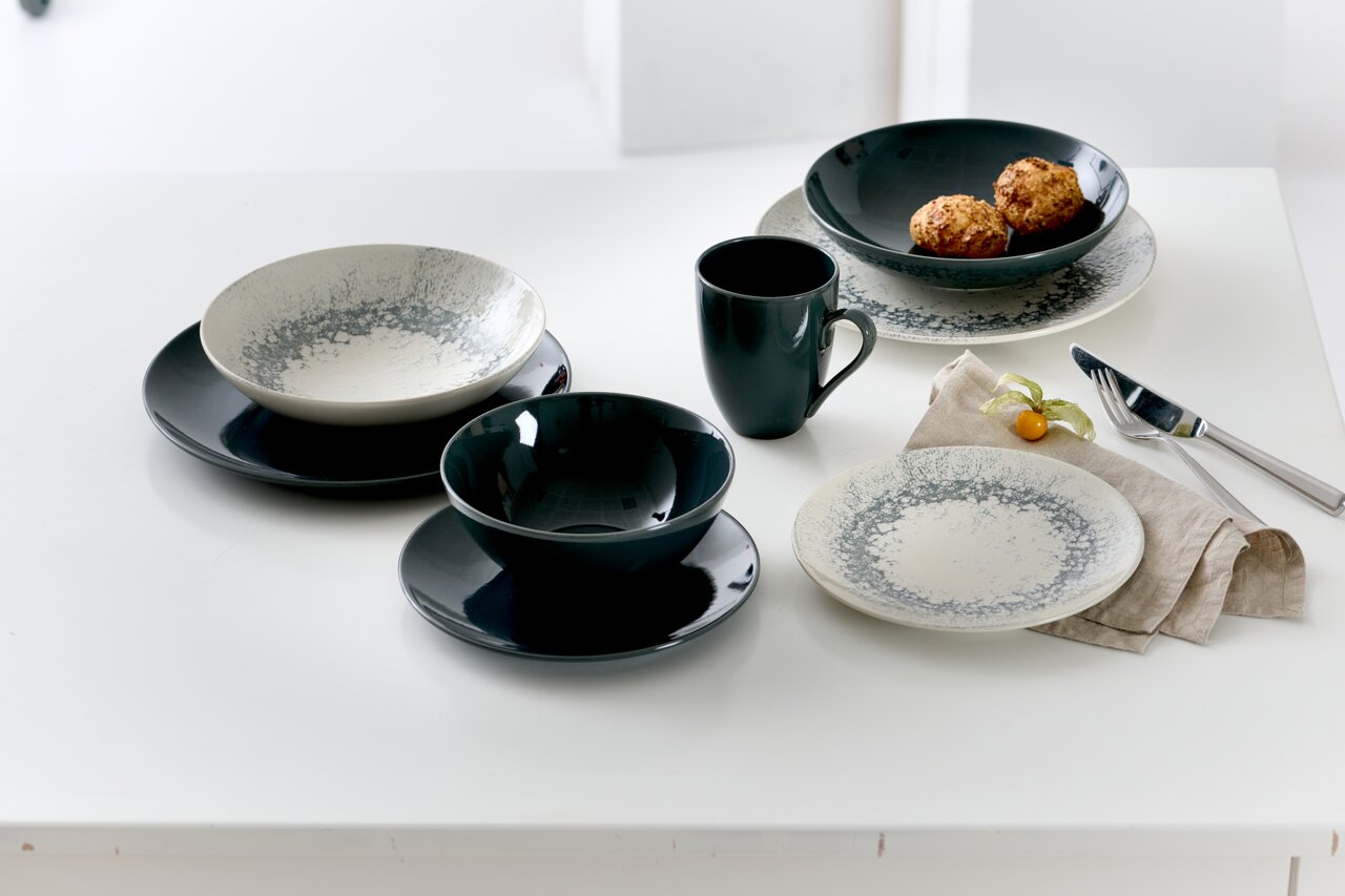 Vogelperspektive auf ein stilvolles Geschirrset mit grauen und cremefarbenen Speisetellern, Schalen und einer Tasse auf einem weißen Tisch.