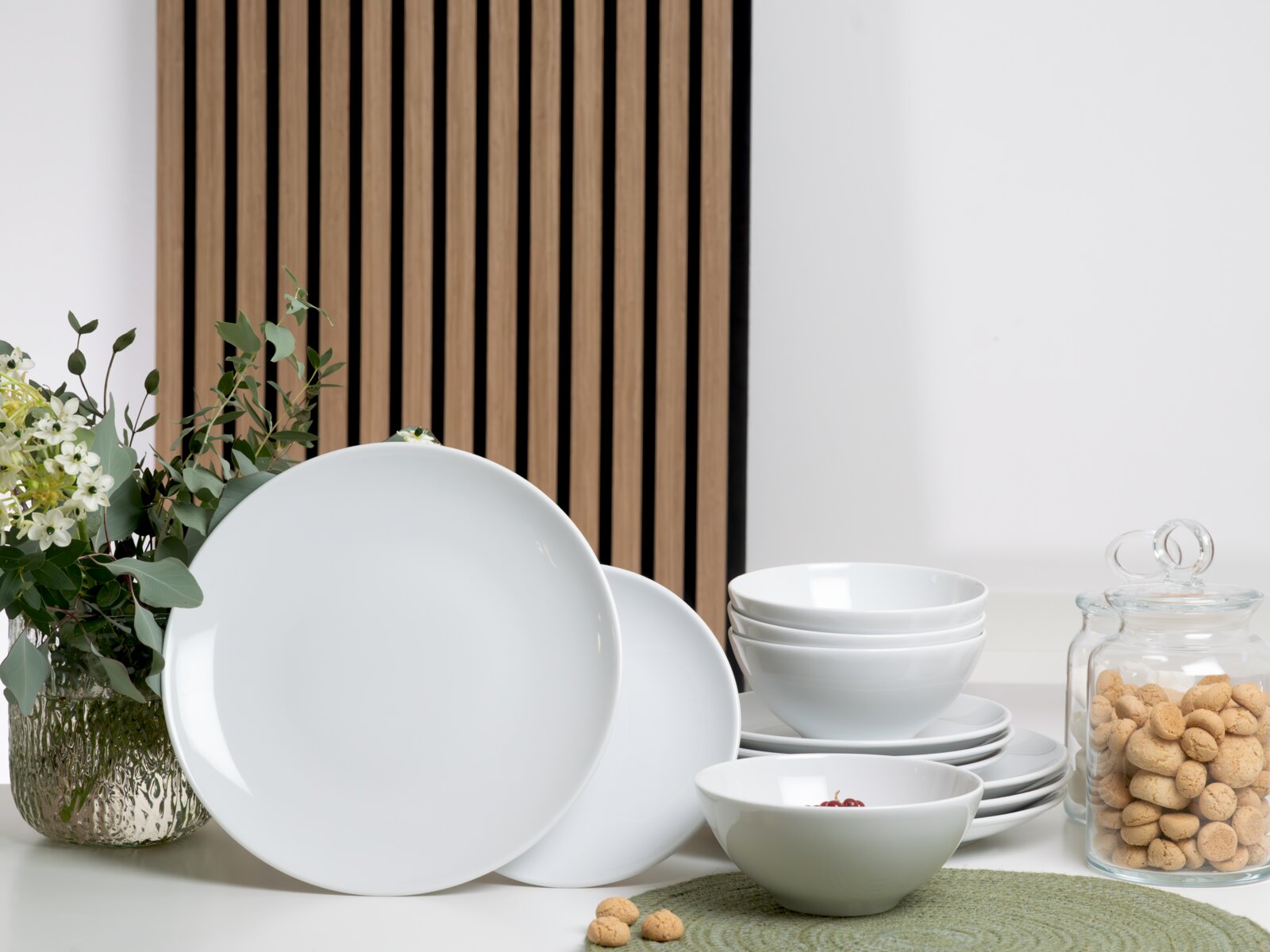 creaTable Tellerset 12-tlg BISTROT Weißes Porzellan-Geschirrset mit großen Tellern, Schalen und kleinen Tellern auf einem Tisch arrangiert, frontal in leichter Seitenansicht fotografiert; daneben Glasdose mit Keksen und Blumenvase vor Holzlamellen-Hintergrund.